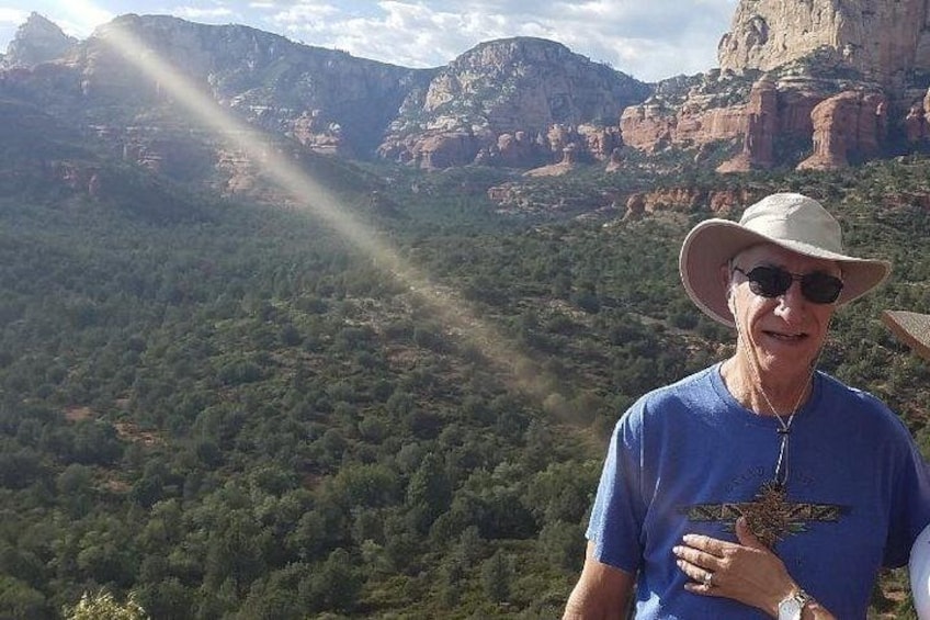 A person wearing a blue shirt and a hat, standing in a mountainous area with greenery and rocky formations. 2