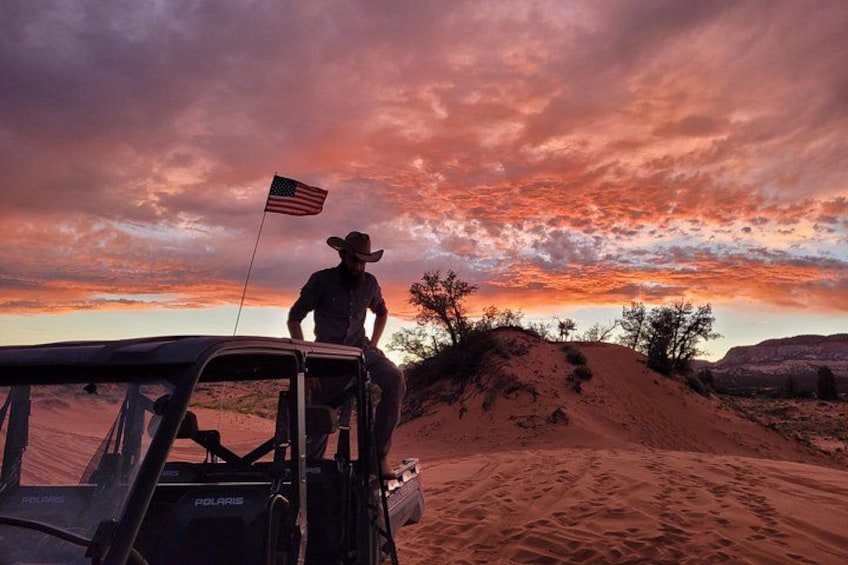 Una persona con sombrero de vaquero sentada en un vehículo en un desierto con dunas de arena y un atardecer. 2