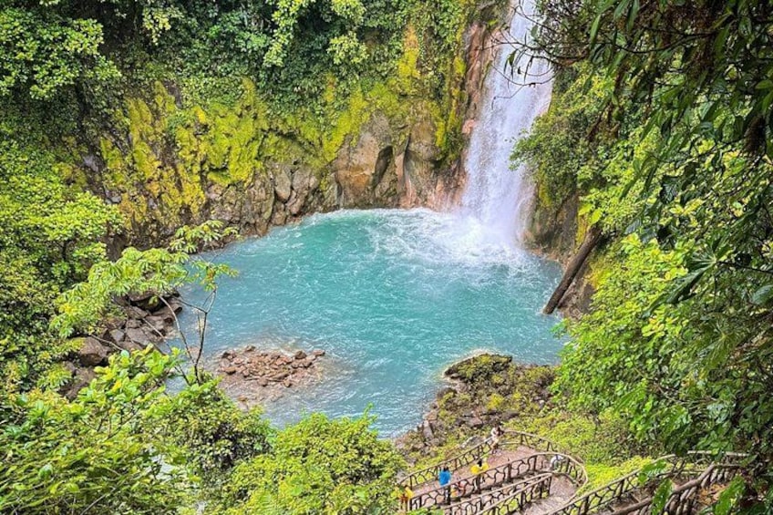 Una cascada, una piscina turquesa y una escalera que desciende hacia la piscina, rodeada de exuberante vegetación. 1