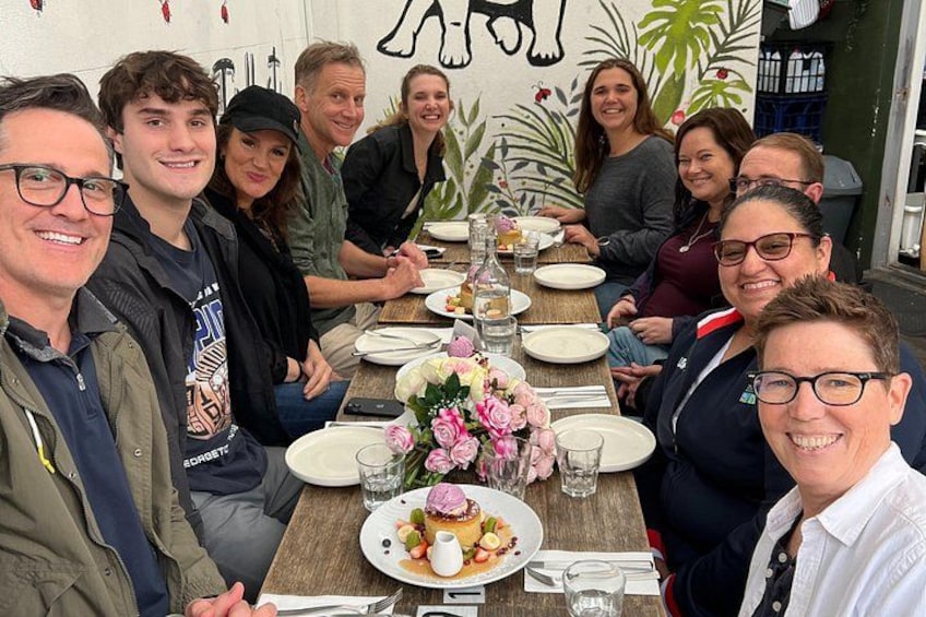Un groupe de personnes est assis autour d’une table en bois, devant des assiettes remplies de nourriture, des verres et un vase de fleurs. 1