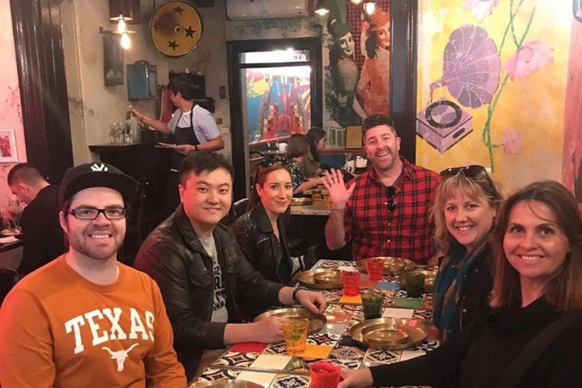 Un groupe de personnes est assis à une table dans un restaurant, devant une fresque murale aux couleurs vives, et on aperçoit un barman au fond. 3