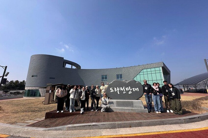 A group of people standing in front of a modern building with a curved facade and a large sign with Korean text. 5