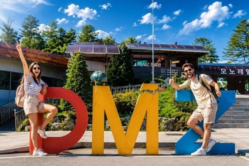Dos personas posan junto a letras grandes 'A' y 'M' en un parque, con un edificio al fondo. 1