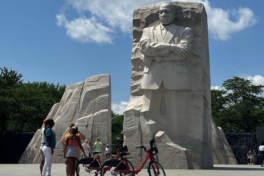 A large stone monument with a seated figure, surrounded by people and bicycles. 4