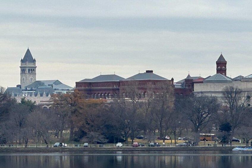 A large building with a clock tower, a body of water, and trees in the foreground. 2