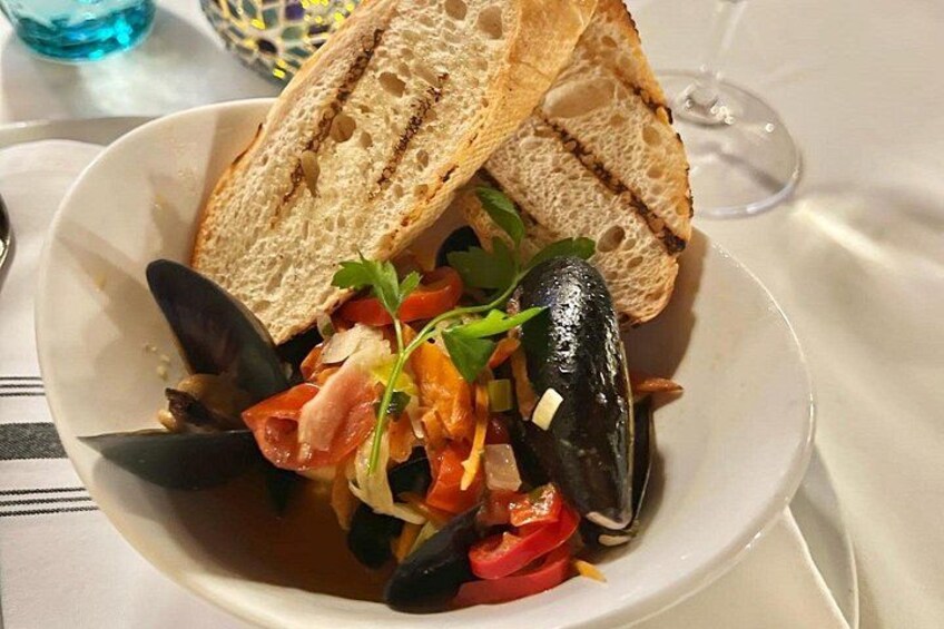 Un plat de fruits de mer composé de moules, de tomates et d’herbes aromatiques, servi dans une assiette blanche, avec un verre de vin en arrière-plan. 2