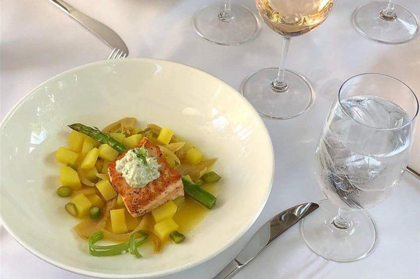 Een wit bord met vis, asperges en gele groenten, samen met wijnglazen en een vork op een tafel. 4