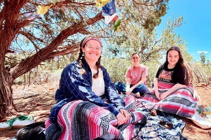 Three people sitting on a blanket under a tree with a desert landscape in the background. 2