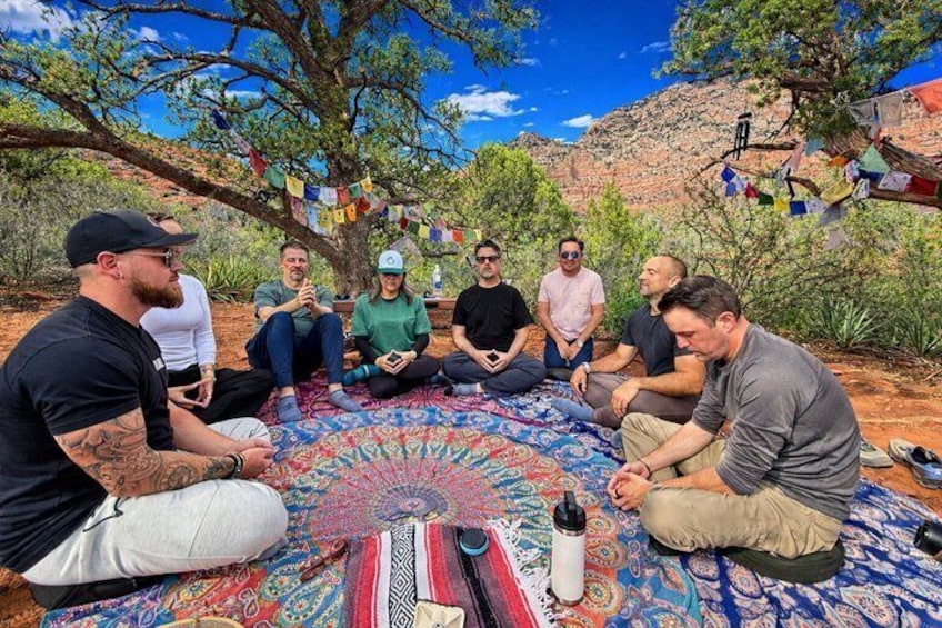 A group of people sitting on a patterned rug outdoors, with trees and a clear sky in the background. 2