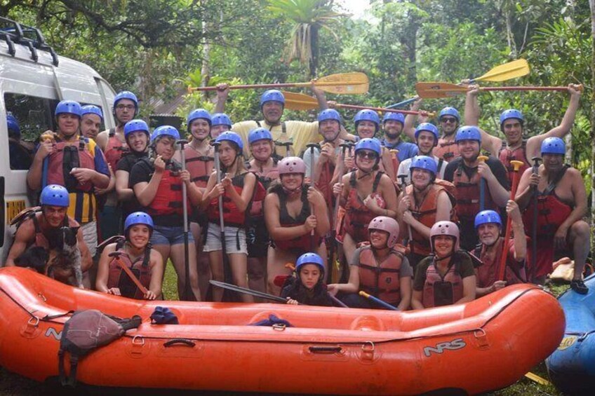 A group of people in life jackets and helmets on a raft, with a vehicle and trees in the background. 1