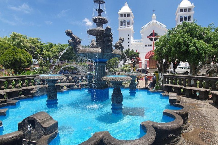 A fountain with multiple tiers and water jets, surrounded by stone structures, with a church and bell towers in the background. 2