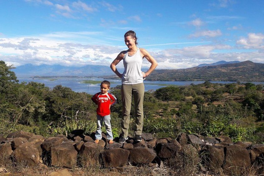 A woman and a child standing on a rocky outcrop with a scenic view of mountains and a lake in the background. 2