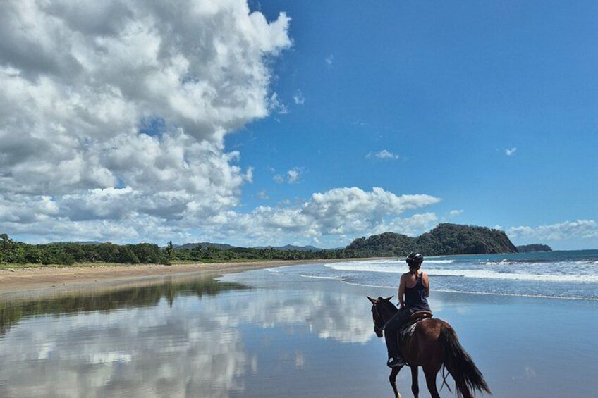 A person riding a horse on a beach with calm water, a distant island, and a partly cloudy sky. 1