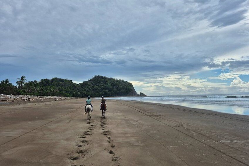 A beach with footprints leading to the water, a horse, and two riders. 2