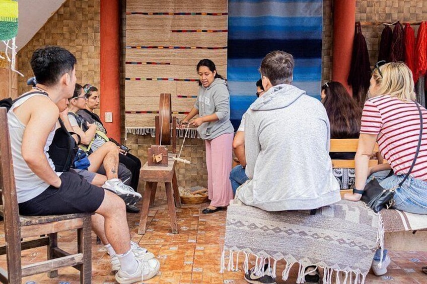 A group of people sitting on wooden stools, engaged in a weaving activity with a loom in a rustic setting. 2