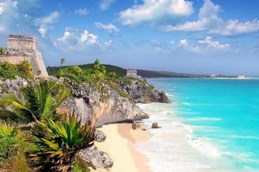 A coastal landscape with a sandy beach, clear turquoise waters, and a historical stone structure. 4