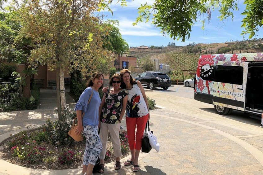 Three people standing near a tree, a bus, and a building in a sunny outdoor setting. 3