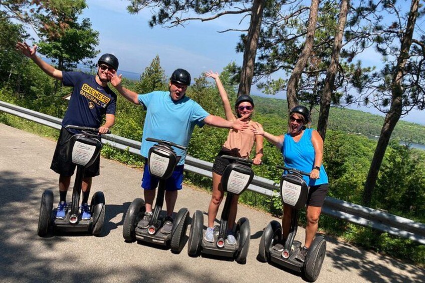 Four people on Segway devices, standing on a road with a railing, surrounded by trees and a view of the landscape. 5