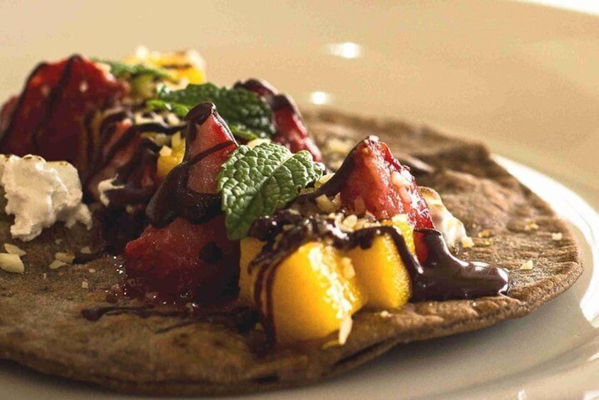 Un plato de postre con un crepe cubierto de chocolate, fresas, mango, hojas de menta y cucharadas de crema. 1