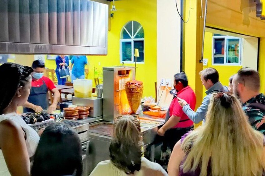 A food preparation area with a large vertical cooking apparatus, a group of people, and a yellow wall. 2