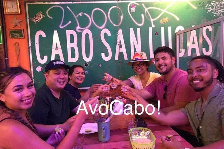 En grupp människor skålar med drycker vid ett bord där det står ett skylt med texten ”Cabo San Lucas”. 1