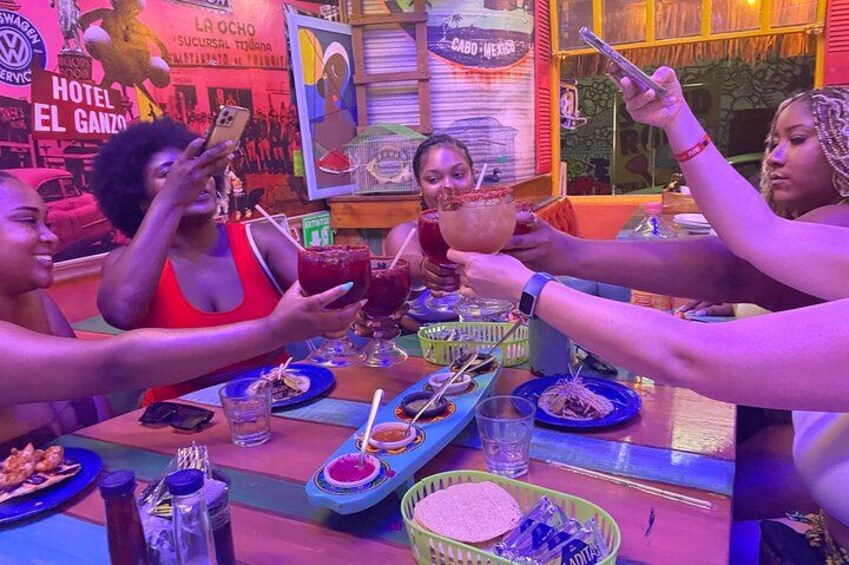 A group of people toasting with drinks at a table with food and a colorful background. 4