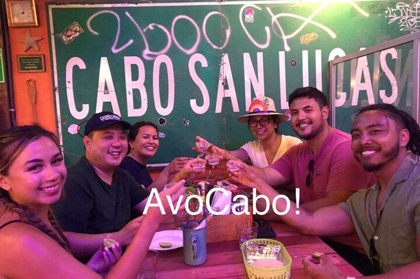 A group of people toasting with drinks at a table with a sign reading 'Cabo San Lucas' in the background. 2