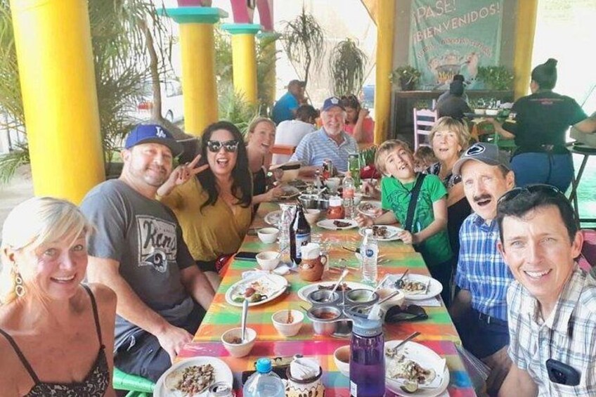 Un grupo de personas cenando en un restaurante al aire libre con un mantel colorido y pilares amarillos. 4