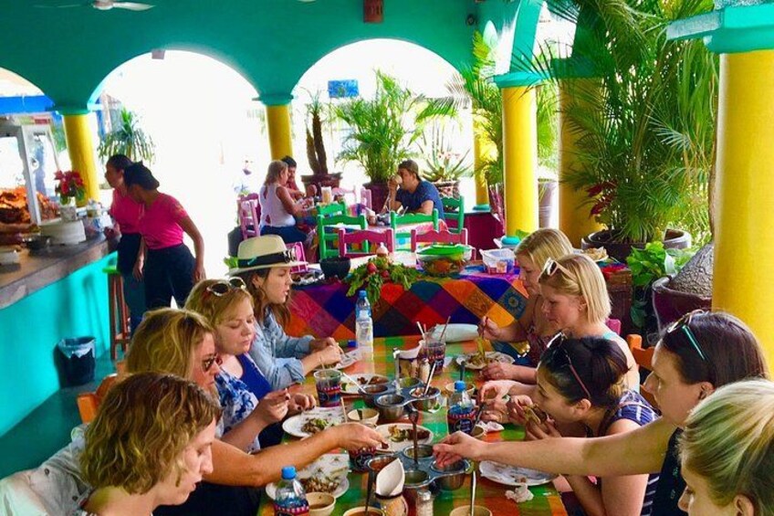 Un grupo de personas cenando en un restaurante al aire libre con mesas coloridas y plantas tropicales. 2