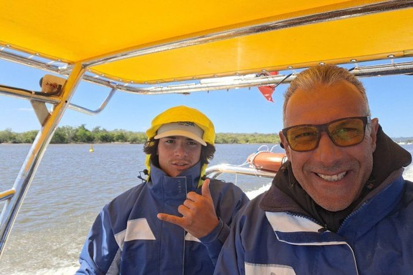 Twee personen in een boot met een geel dak, water op de achtergrond en begroeiing langs de oever. 2