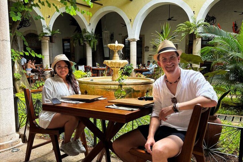 Twee personen zitten aan een houten tafel in een binnenplaats met een fontein op de achtergrond. 3