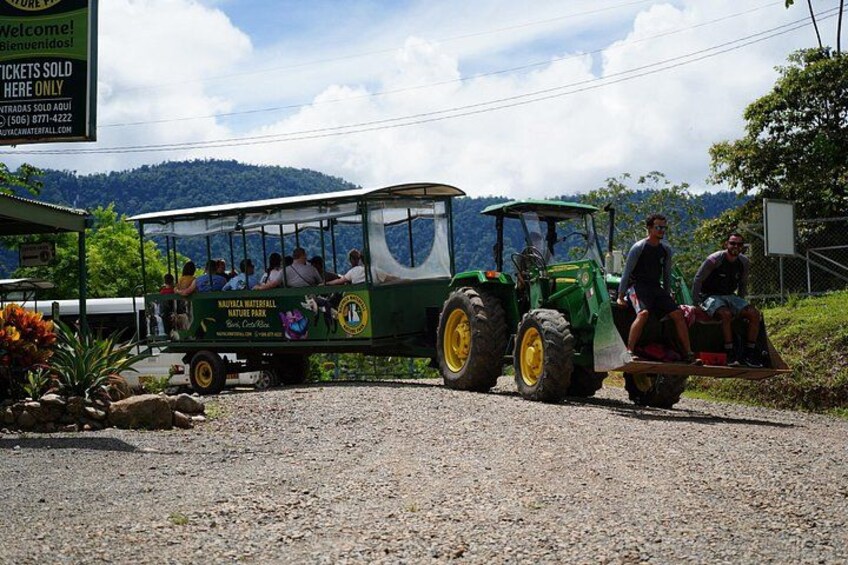 Een groene tractor trekt een passagierstrein met mensen aan boord, geparkeerd op een grindweg met een bergachtig landschap op de achtergrond. 2