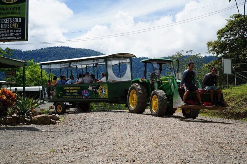 Un tractor verde remolca un carruaje con pasajeros sentados, estacionado en un camino de grava con un paisaje montañoso al fondo. 2