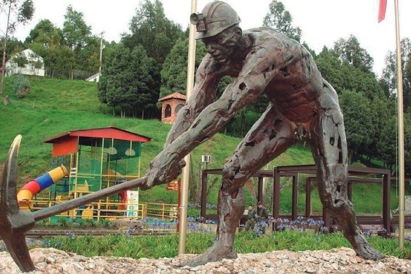 Una estatua de bronce de un hombre con un pico, ubicada en un parque con un área de juegos y árboles al fondo. 2