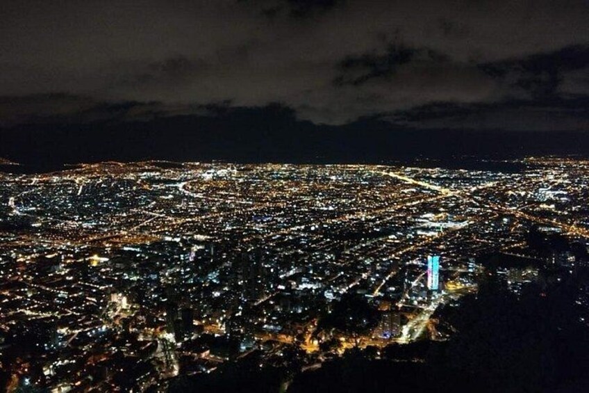 Nachtansicht einer Stadt aus der Vogelperspektive mit dichtem Licht, einem markanten, blau beleuchteten Turm und einem bewölkten Himmel. 3