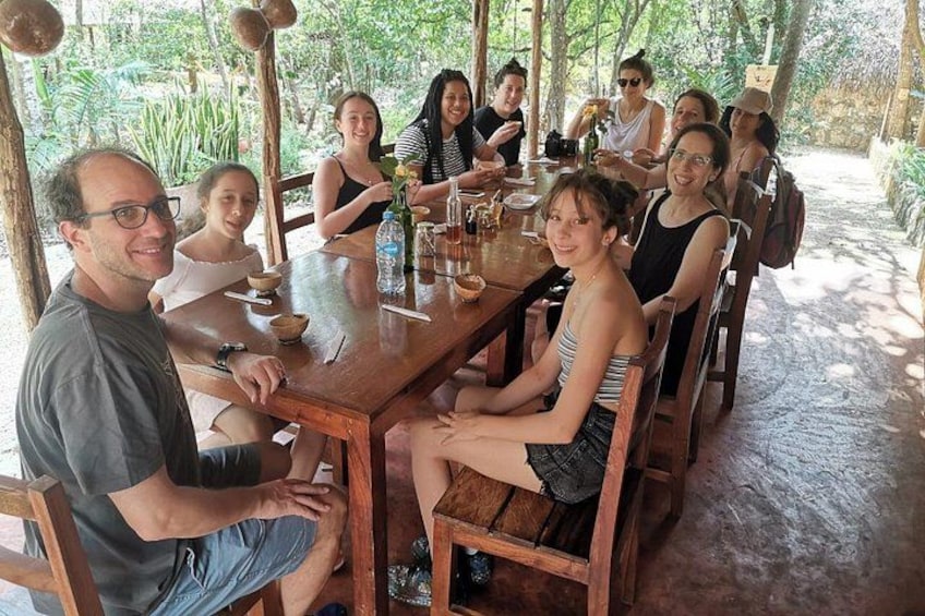 A group of people sitting around a wooden table outdoors, with greenery in the background. 2