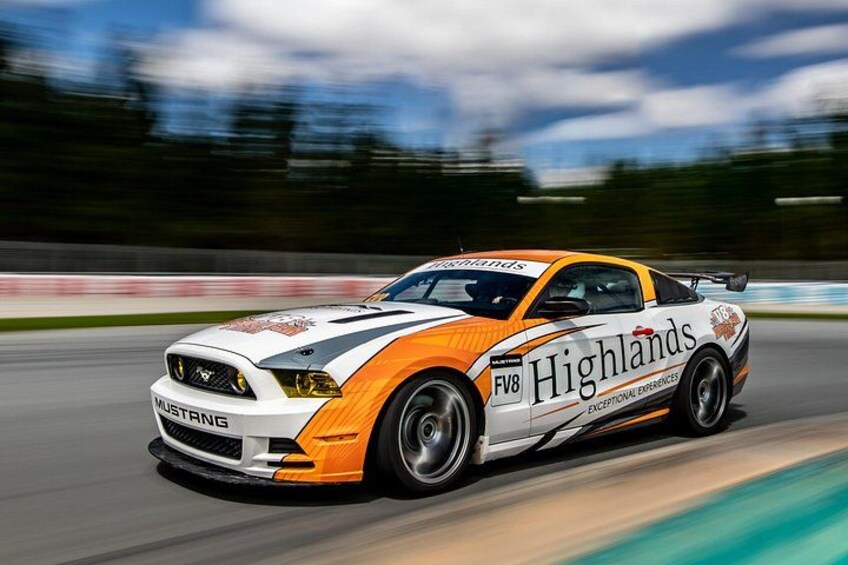 Une voiture de course blanche et orange, avec les mots « Highlands » et « Expériences exceptionnelles » inscrits sur les flancs, circule sur une piste. 1