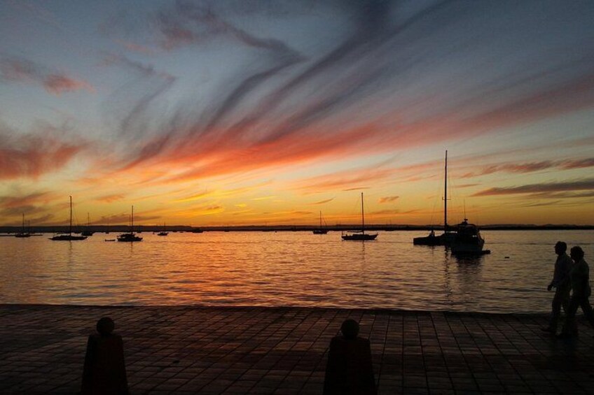 A sunset over a body of water with sailboats and people on a brick-paved area. 2
