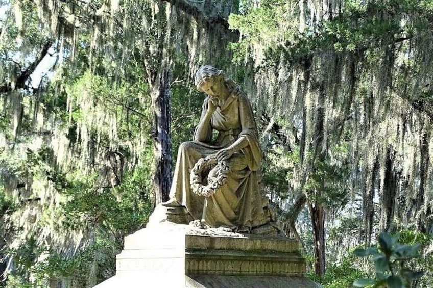 A statue of a woman sitting on a pedestal surrounded by trees and moss. 1