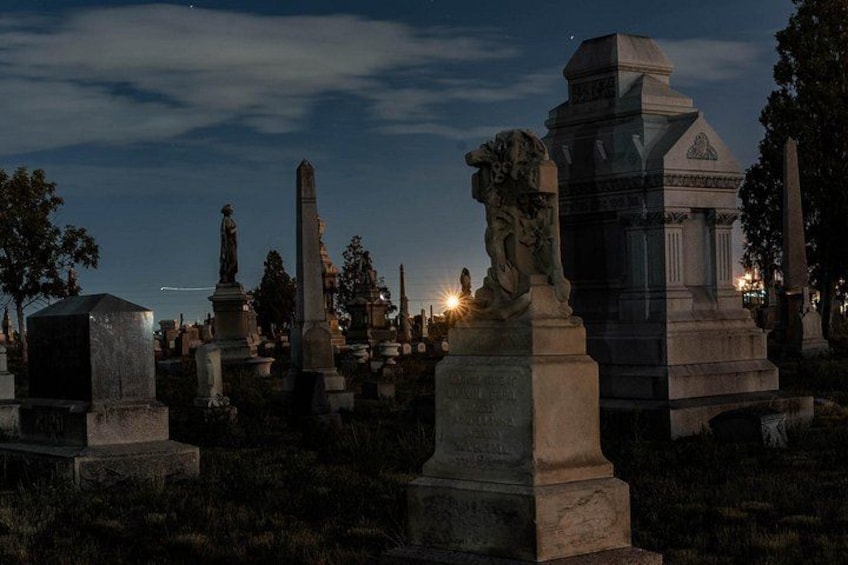 A cemetery with various tombstones and monuments, including a prominent obelisk and a statue. 3
