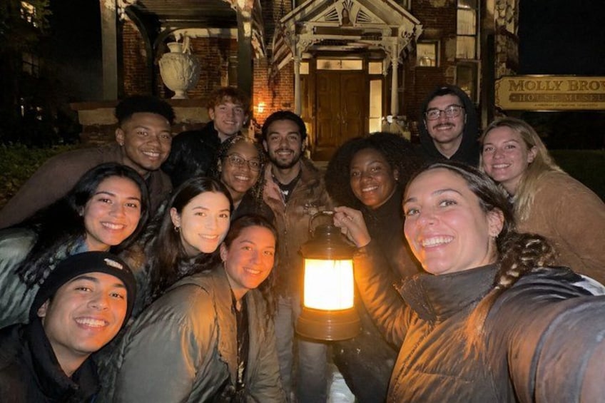A group of people gathered around a lit lantern in front of a building with a sign that reads 'Molly Brown's.' 1