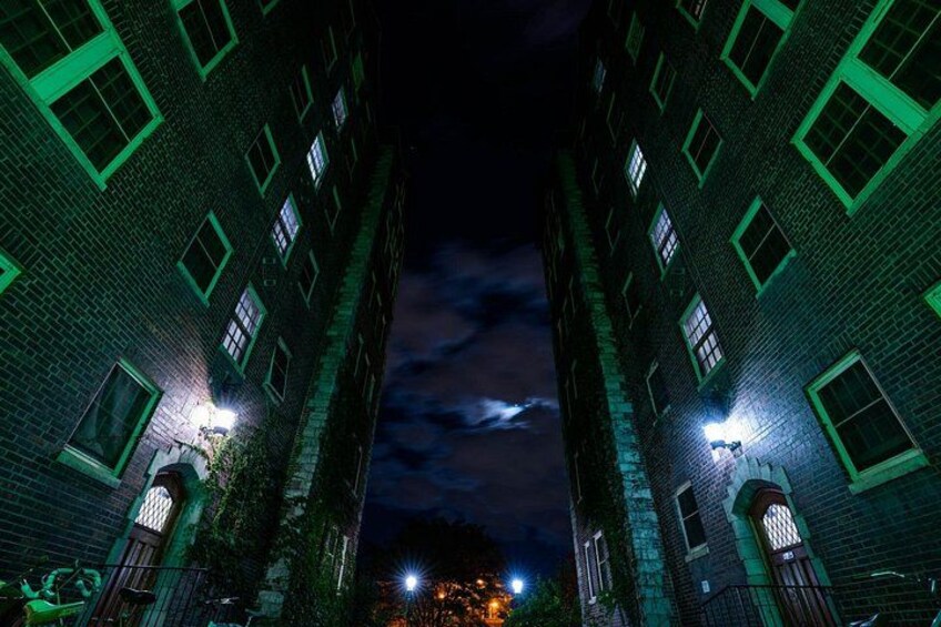 A nighttime view of a narrow alley between two brick buildings with illuminated windows and street lamps. 5