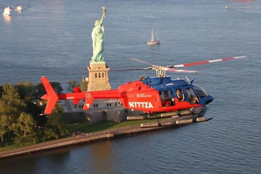 En röd och blå helikopter med texten ”NTTZA” målad på sidan, som flyger över en sjö med Frihetsgudinnan synlig i bakgrunden. 3