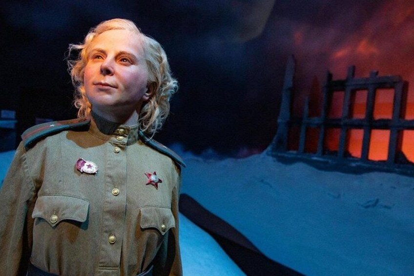 En person i militäruniform, med medaljer på bröstet, som står framför en scen med en mörk bakgrund och ett staket. 2