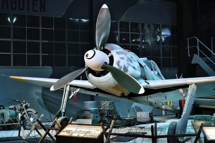 Ett gammalt flygplan visas upp med sin propeller, i en museimiljö med andra utställningsföremål i bakgrunden. 5