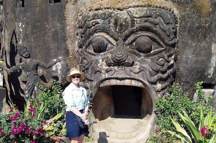 Une personne se tient devant une sculpture représentant un visage en pierre, avec un grand œil et une grande bouche, le tout entouré de verdure et de fleurs. 1