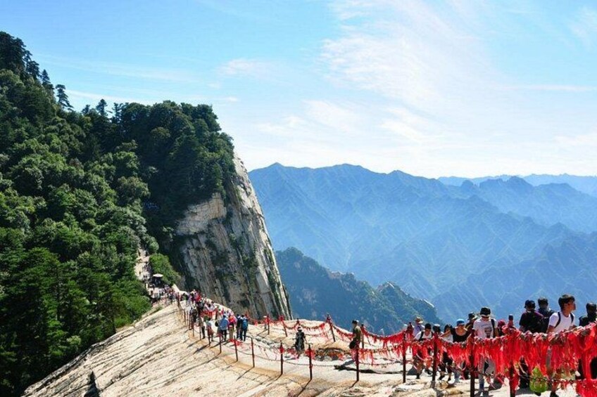 A mountainous landscape with a steep cliff, a group of people on a viewing platform, and a forested area. 2