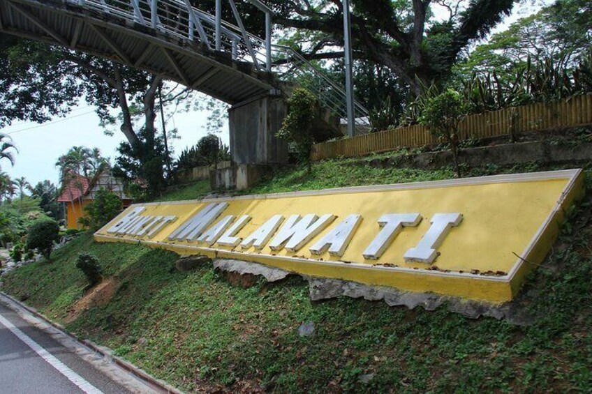 A yellow sign with 'Batu Malawati' written on it, set against a backdrop of greenery and a bridge. 1