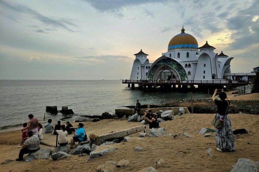 Een strand met mensen die zitten en staan, een groot wit gebouw met een gouden koepel, en een wateroppervlak. 3