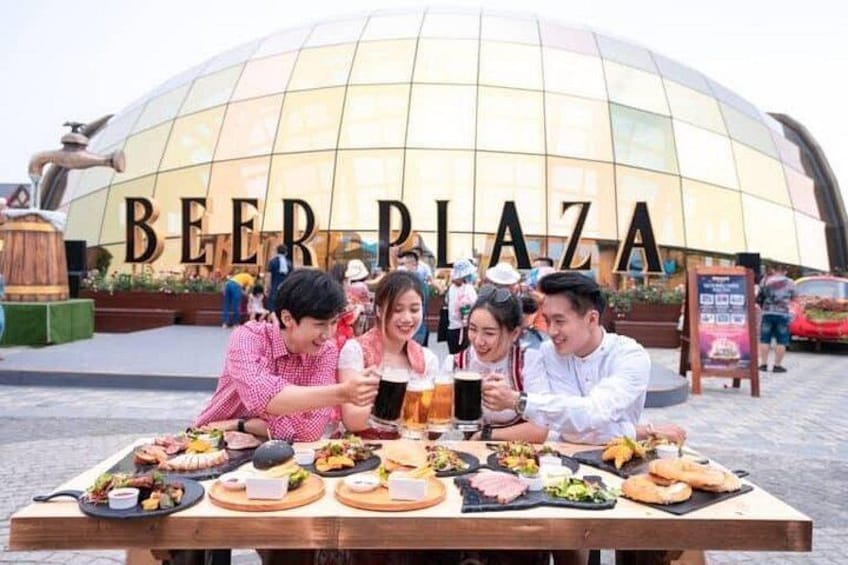 A group of people toasting with beer glasses at a table with various food items in front of a building labeled 'BEER PLAZA.' 2
